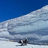 Two skiers in adapted equipment next to the snow wall of the Laguna chairlift, next to the Veleta peak.