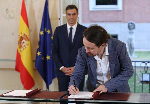 Pablo Iglesias signs the agreement as Pedro Sánchez looks on.