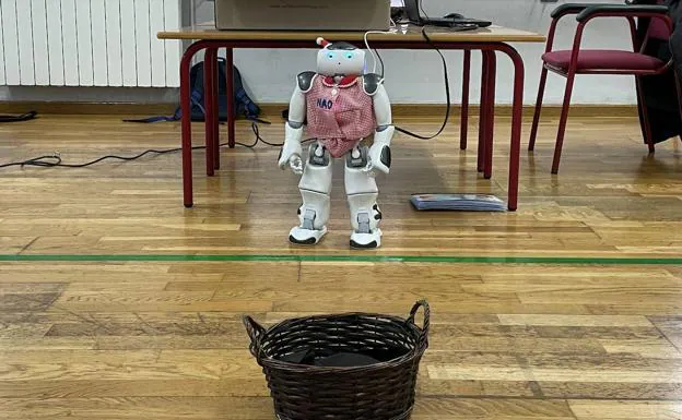 El robot NAO en el aula frente a la cesta donde depositará las pelotas de colores.